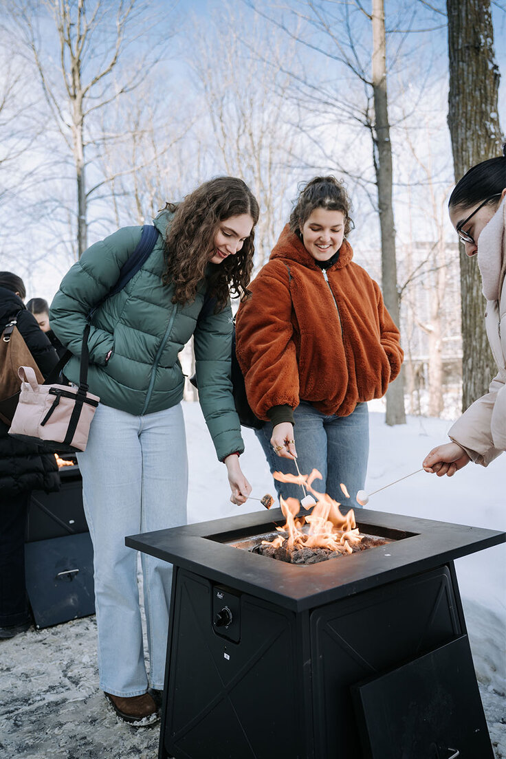 Trois étudiantes de HEC Montréal sont rassemblées à l’extérieur en hiver autour d’un foyer, faisant griller des guimauves et partageant un moment convivial sur le campus.