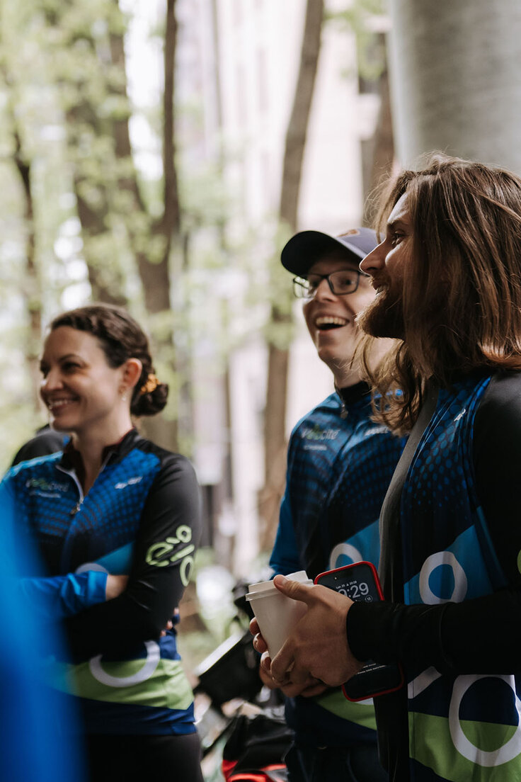 Trois personnes souriantes en tenue de cyclisme  à l’extérieur près d’un bâtiment, l’une tenant une tasse et un téléphone.