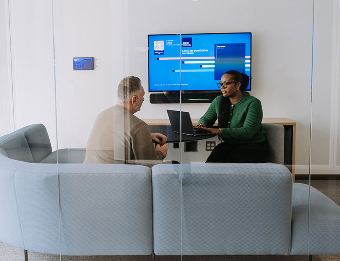Deux personnes en discussion autour d’un ordinateur portable dans une salle de réunion, avec un écran affichant une interface universitaire. 