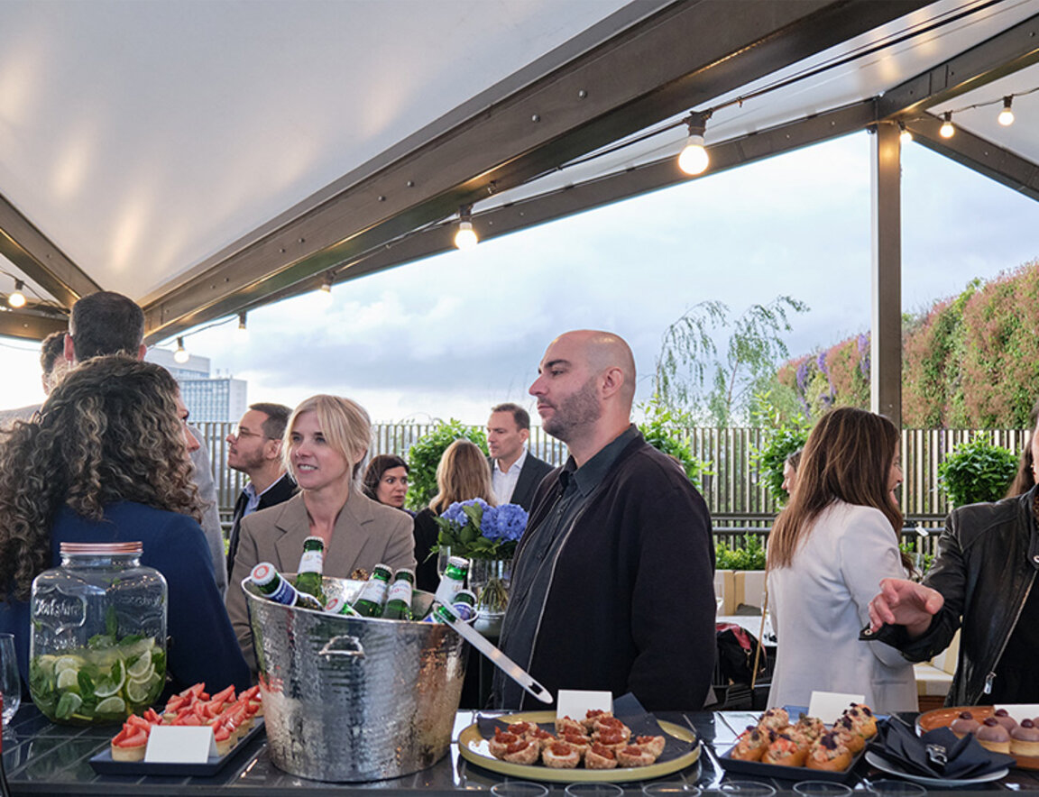 Quelques personnes participent à un cocktail autour d’un bar sur la terrasse d’un édifice.