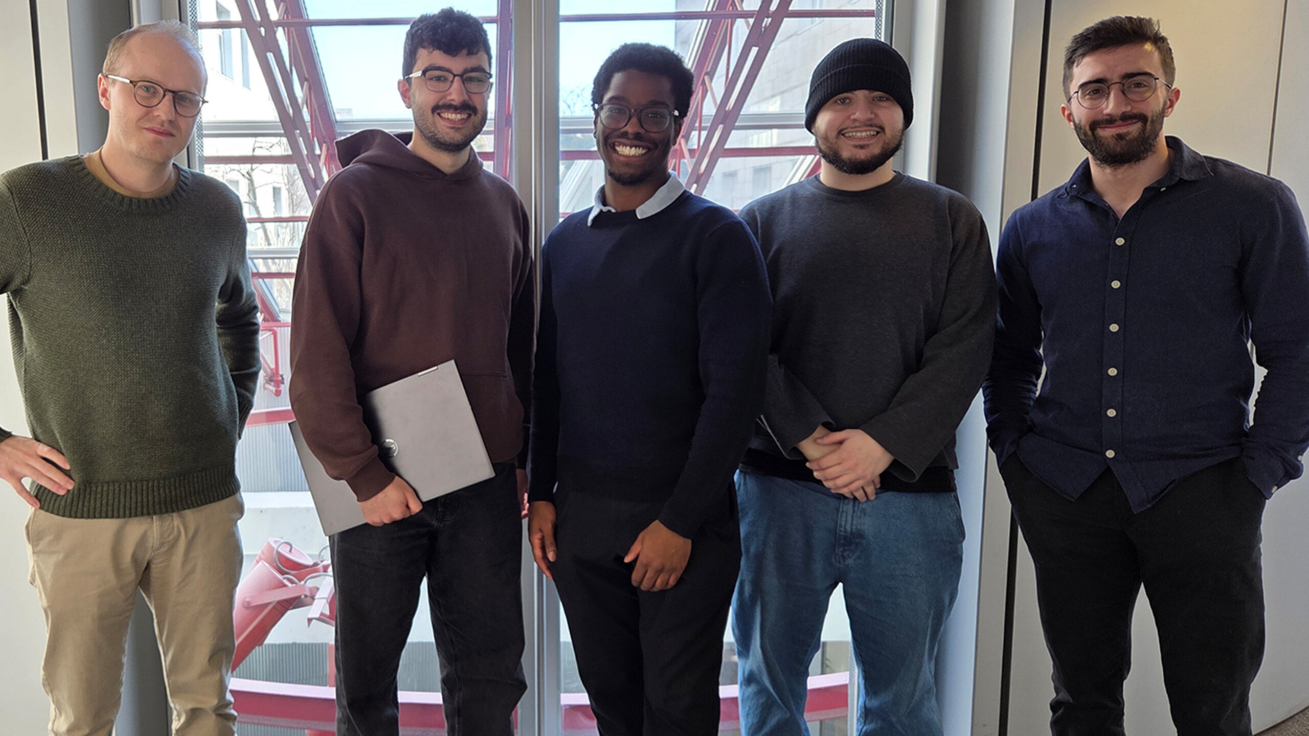 Cinq personnes souriantes debout côte à côte dans un espace intérieur lumineux, devant de grandes fenêtres.