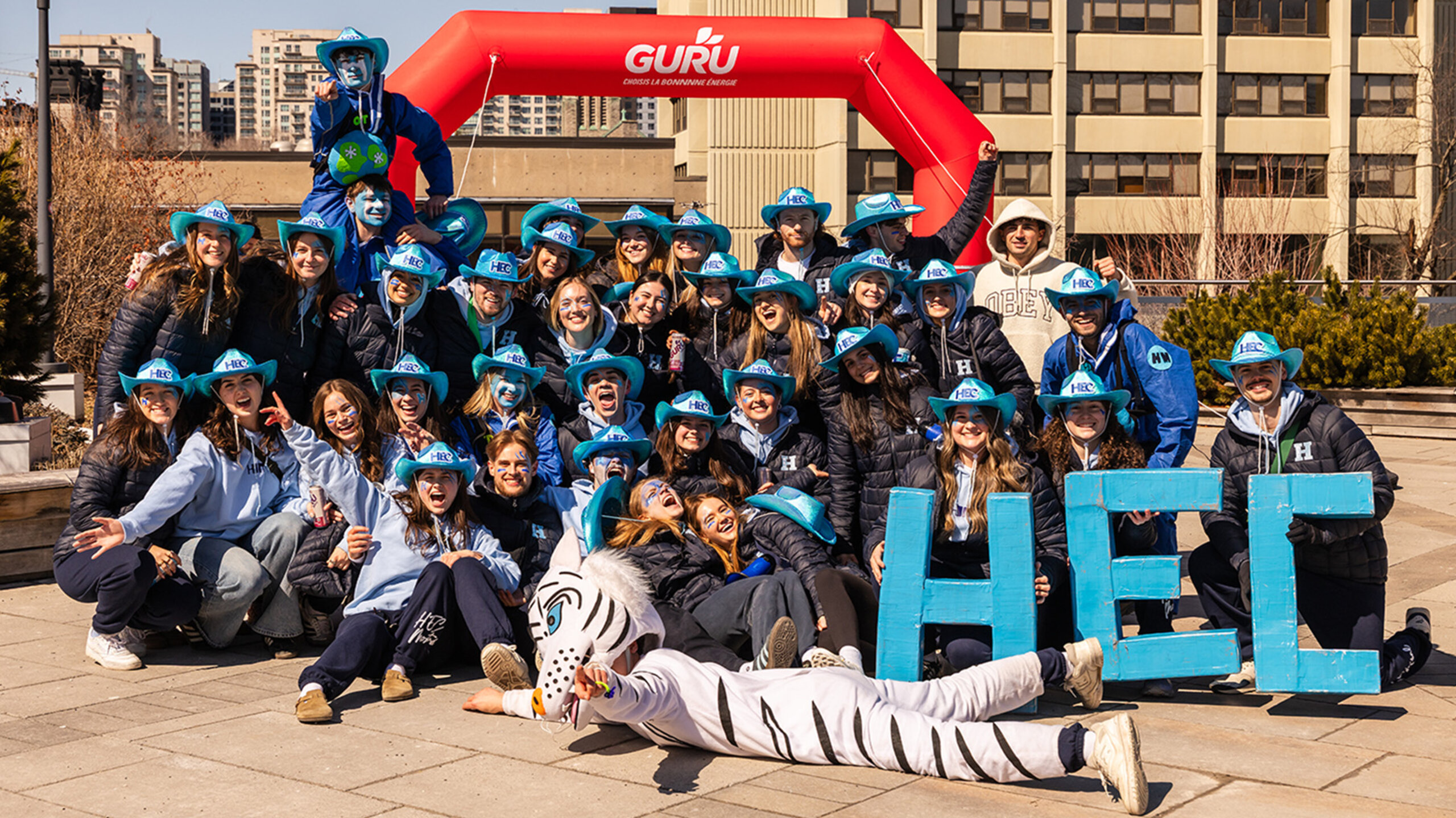 Groupe de la communauté HEC Montréal réuni à l’extérieur, devant une arche événementielle rouge « GURU », avec les lettres « HEC » au premier plan et une mascotte, sur un campus urbain.