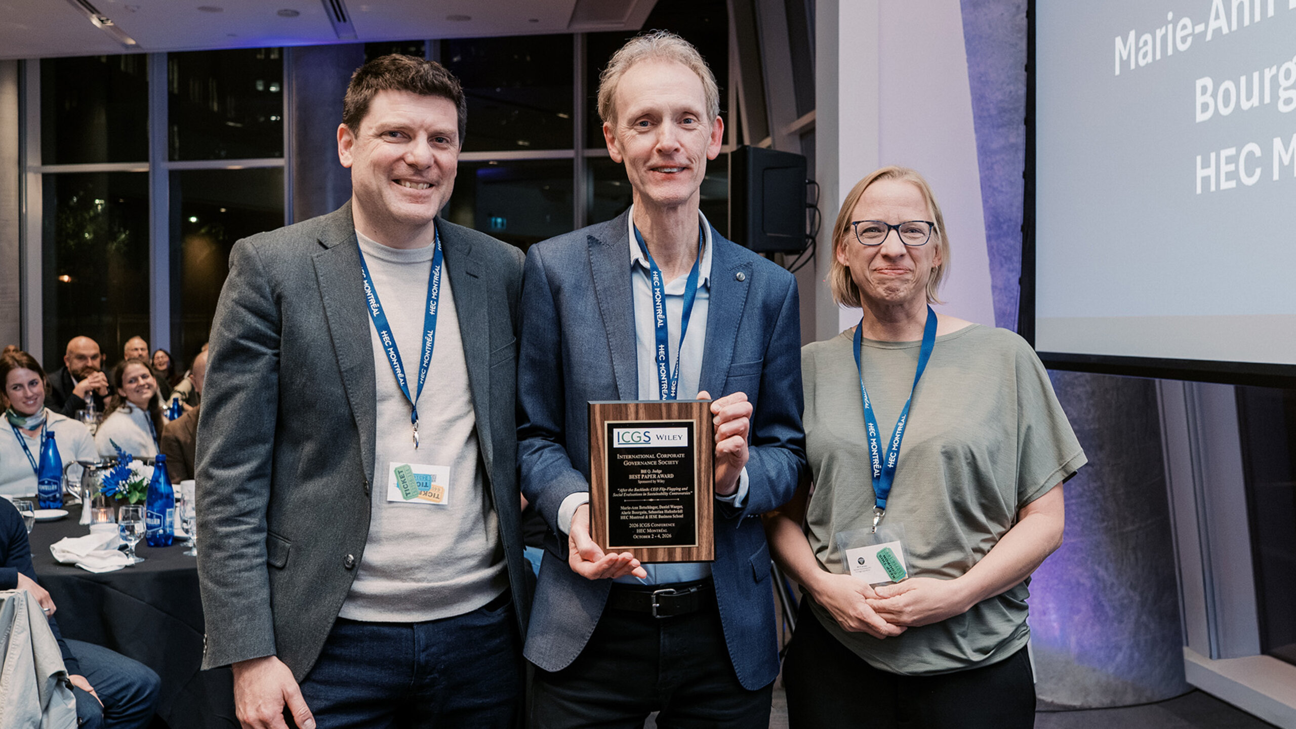 Daniel Waeger, Niels Hermes (ICGS) et Marie-Ann Betschinger posent avec une plaque commémorative devant les participants réunis dans la salle.