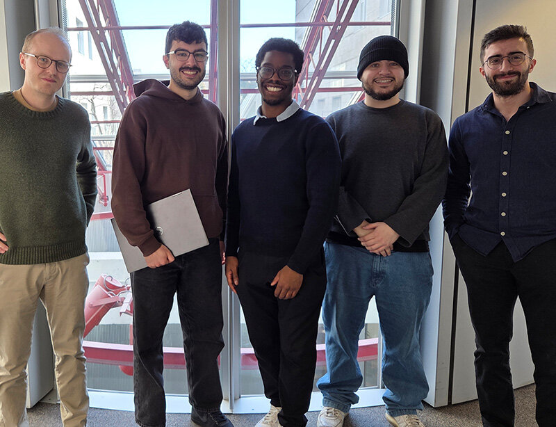 Cinq personnes souriantes debout côte à côte dans un espace intérieur lumineux, devant de grandes fenêtres.
