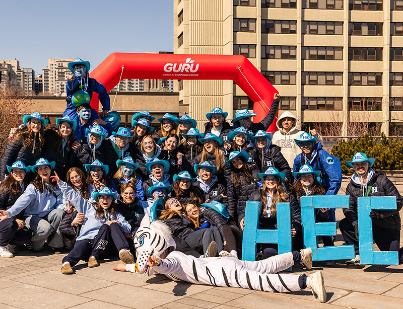 Groupe de la communauté HEC Montréal réuni à l’extérieur, devant une arche événementielle rouge « GURU », avec les lettres « HEC » au premier plan et une mascotte, sur un campus urbain.