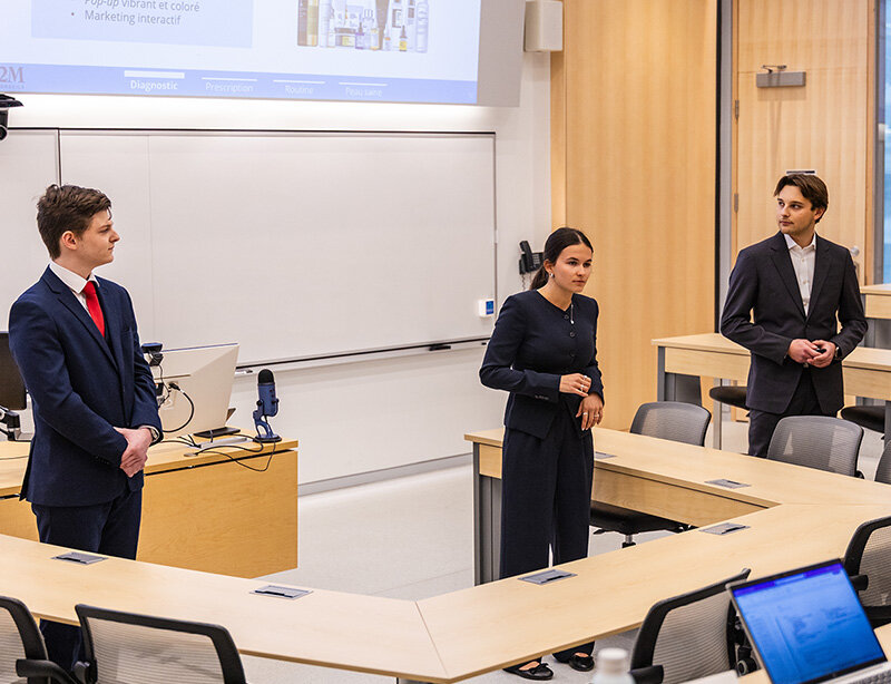 Three people are standing in front of a classroom at HEC Montréal, giving a presentation.