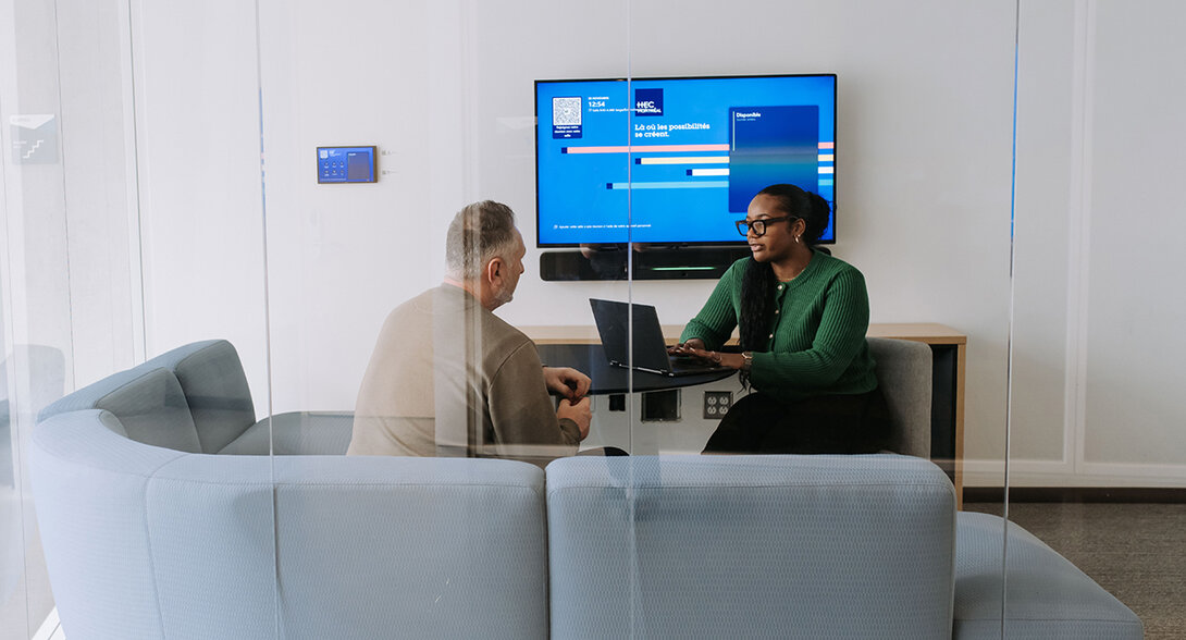 Deux personnes en discussion autour d’un ordinateur portable dans une salle de réunion, avec un écran affichant une interface universitaire. 