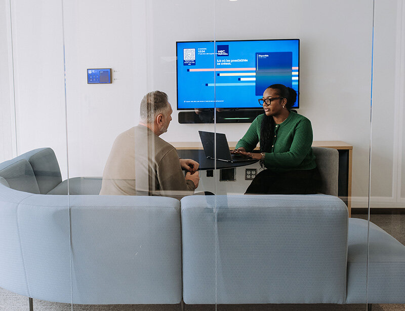 Deux personnes en discussion autour d’un ordinateur portable dans une salle de réunion, avec un écran affichant une interface universitaire. 