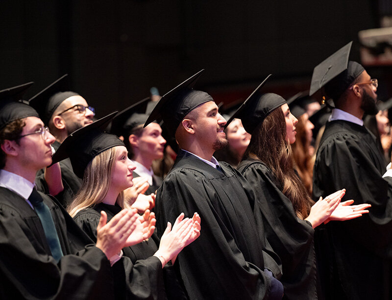 Plusieurs étudiantes et étudiants de HEC Montréal vêtus de toges noires, se tenant debout dans les gradins d’une salle de spectacle, lancent leur mortier dans les airs.